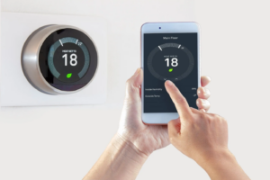 Homeowner using smart thermostat and holding up thermostat app on smartphone to show setting the temperature via app.